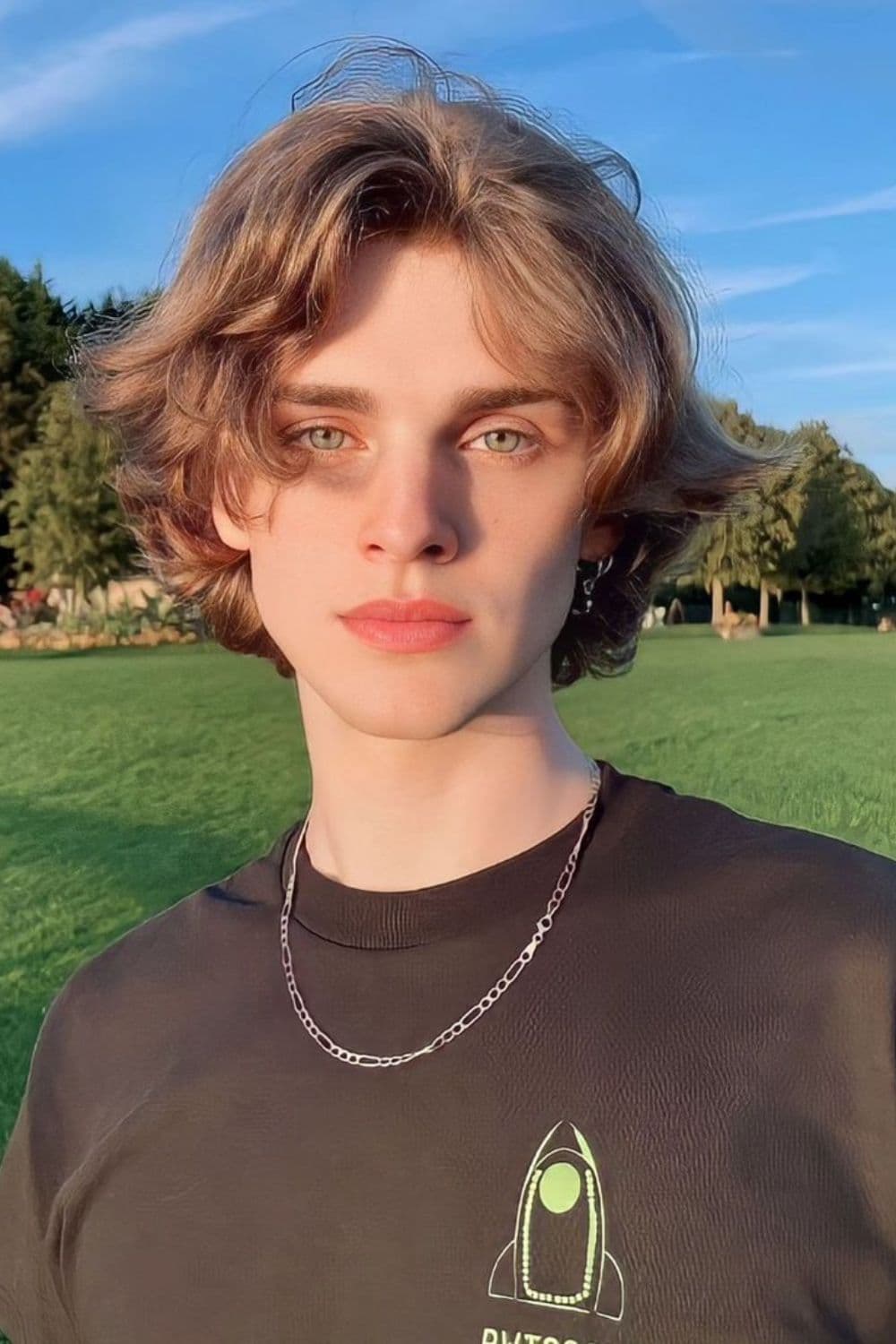 A man wearing a black t-shirt and a silver necklace with blonde 90s/eBoy wolf cut.