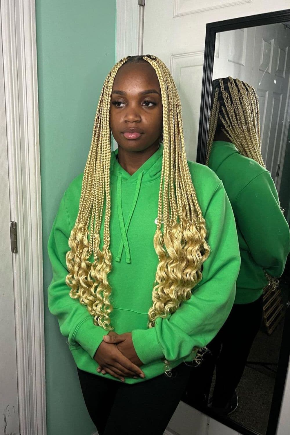 A woman standing wearing a green sweater with blonde medium knotless braids with curly ends.