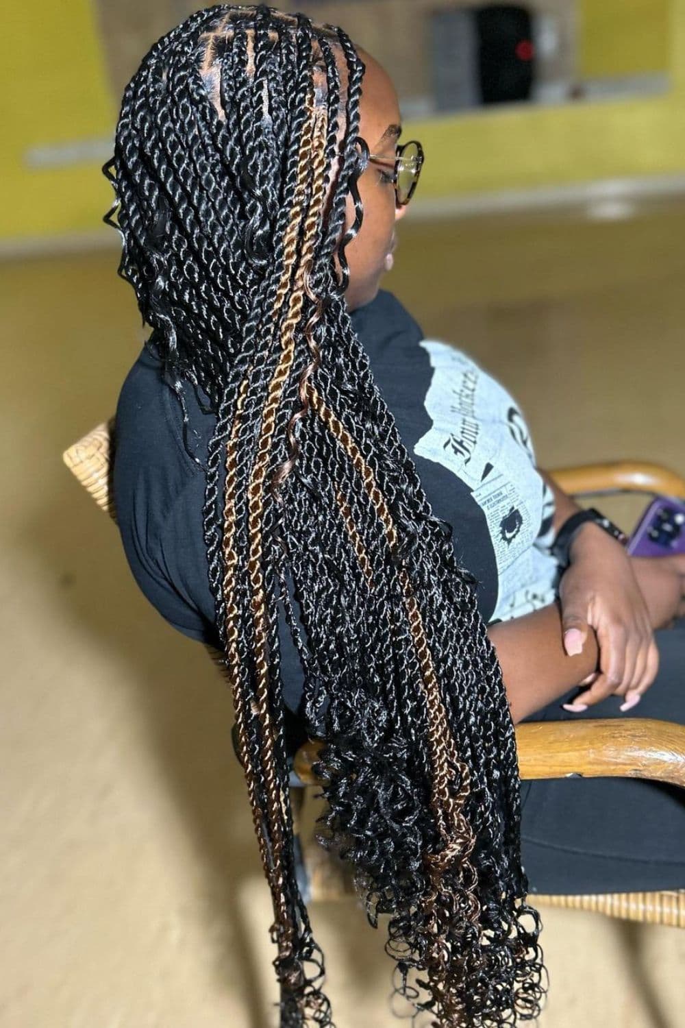 Side view of a woman sitting on a chair with black and brown cornrow braids with curly ends.