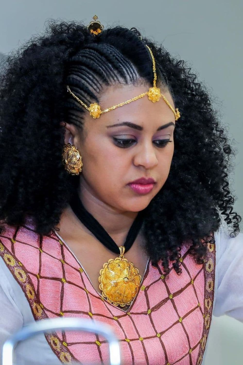 A woman with cornrows with gold hair accessory, gold earrings and gold necklace.