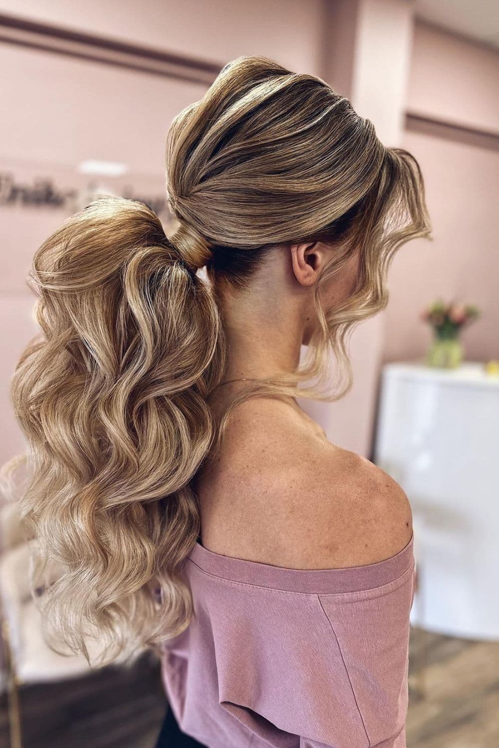 Side view of a woman in pink off shoulder with a blonde curly low ponytail.