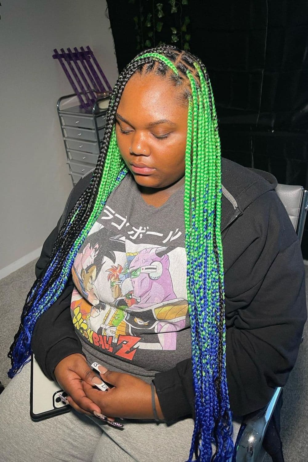 A woman sitting in a salon with green and blue knotless braids.