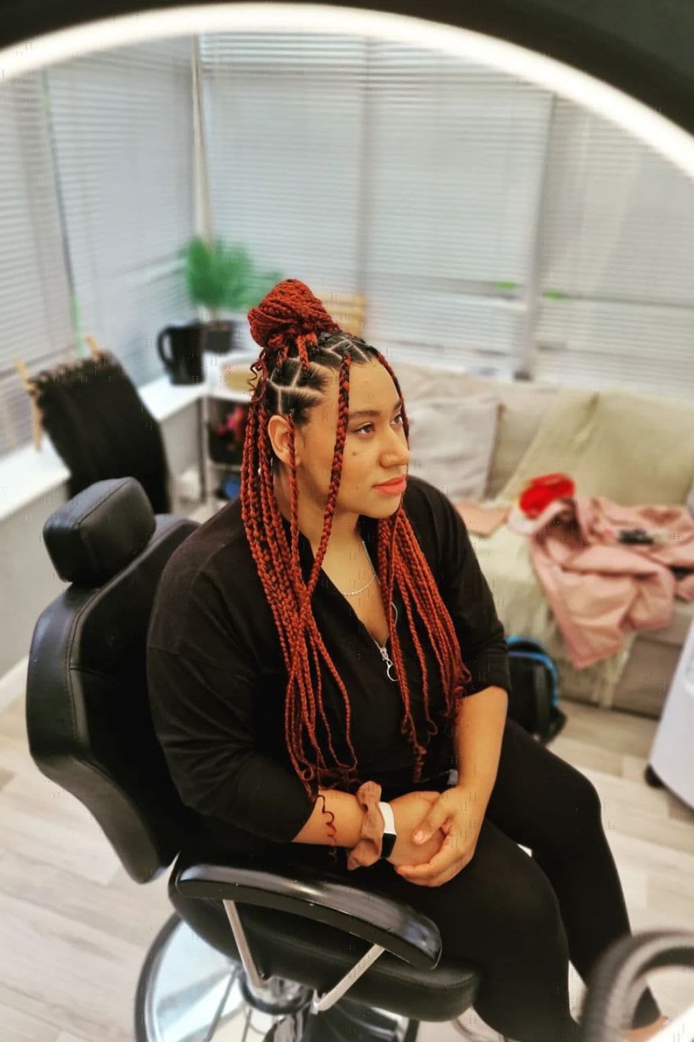 Side view of a woman sitting on a chair in a salon with red half up half down box braids with curly ends.