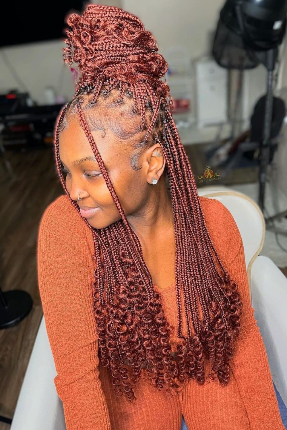 A woman in an orange long sleeves blouse with burgundy half up half down medium knotless braids with curly ends.