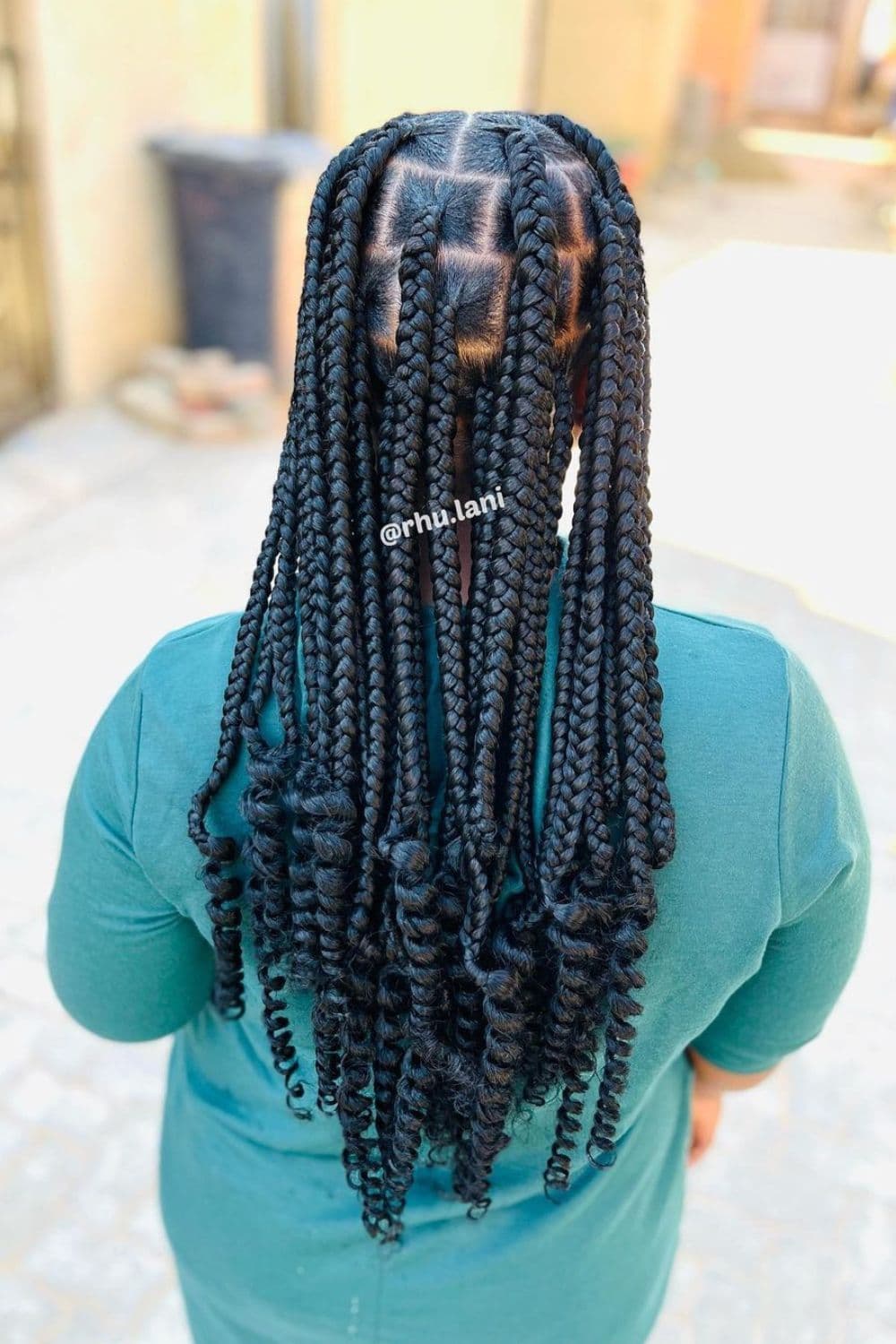 A woman in blue-green long sleeves with black jumbo box braids with curly ends.