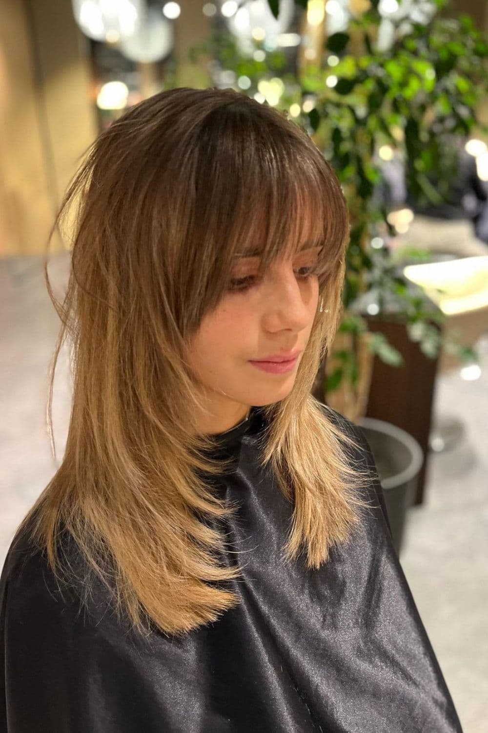 A woman sitting in a hair salon with a long, blonde front layers with classic bangs.