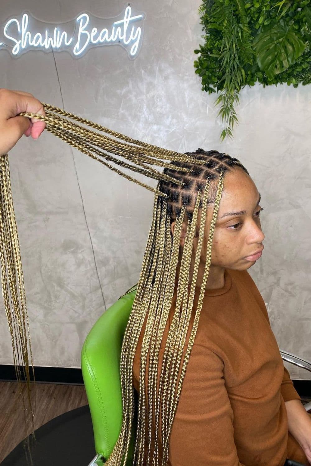 A woman sitting in a hair salon with mixed blonde knotless braids with black roots and a person's hand holding her hair.
