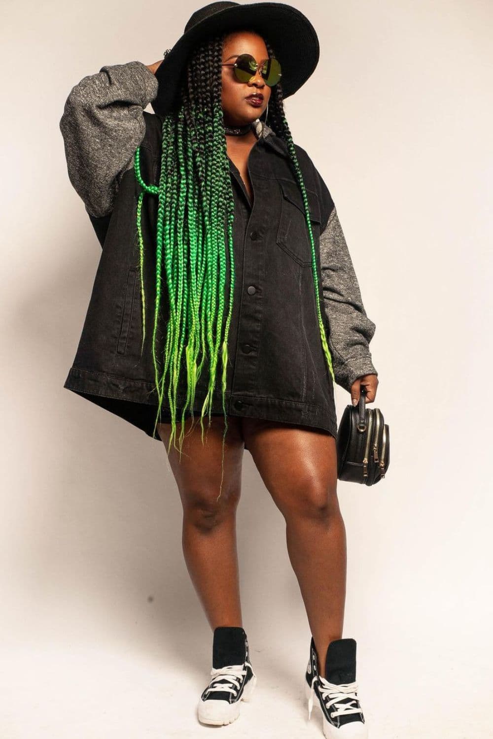 A woman wearing a black and gray jacket, a black hat, sunglasses, black Converge shoes and with shades of green knotless braids.