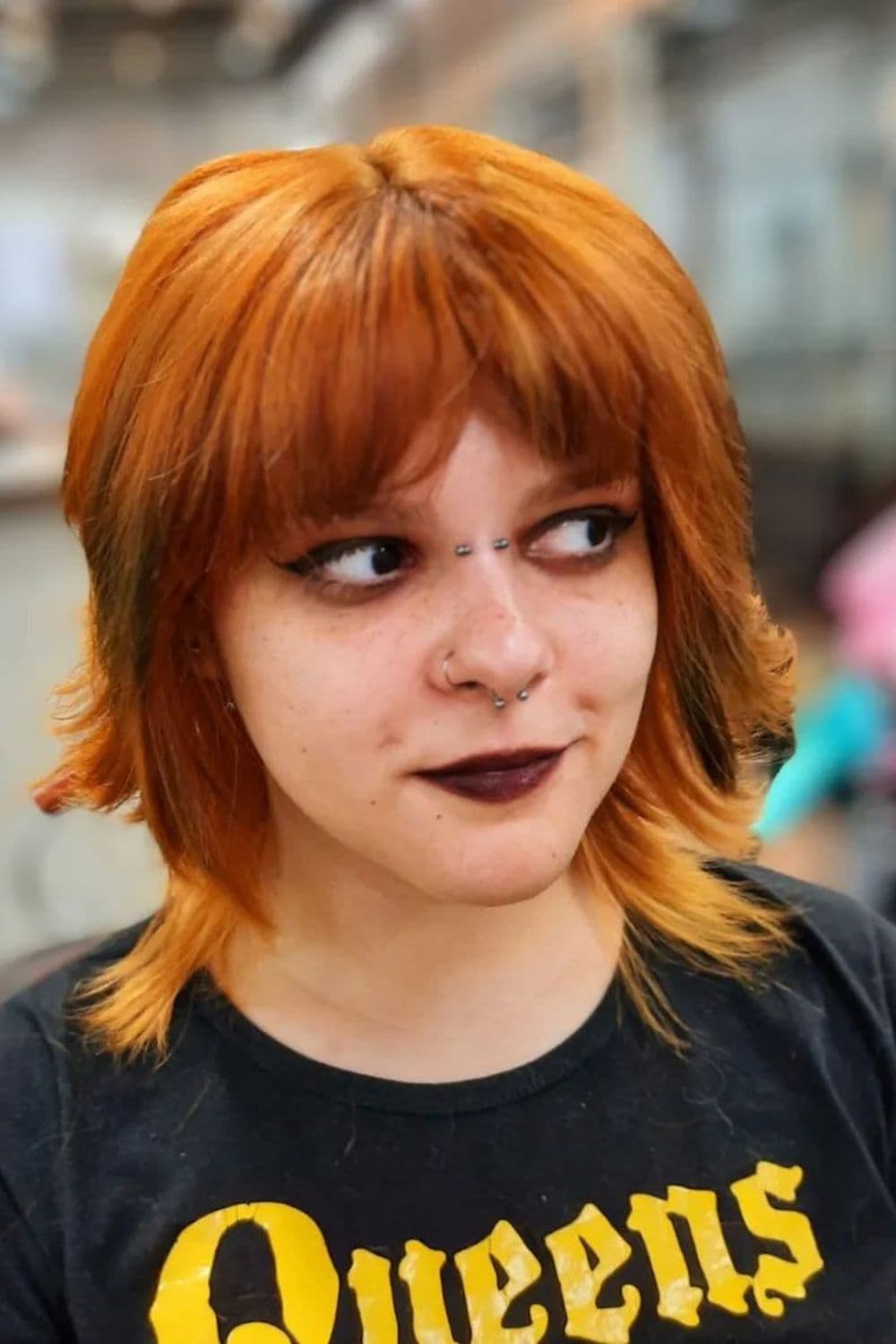 A woman wearing black lipstick and nose earrings with an orange shaggy short wolf cut.