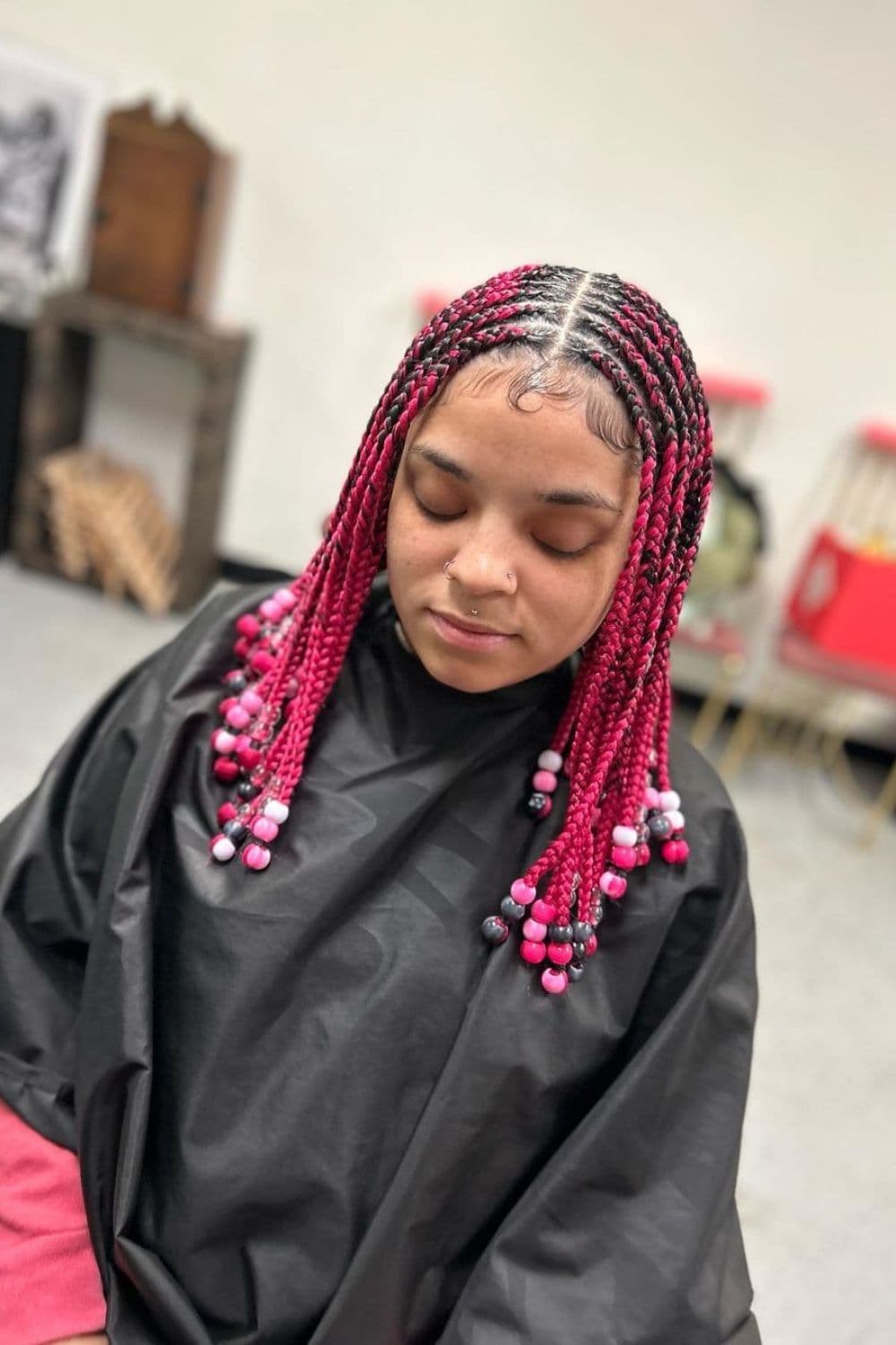 A woman sitting in a hair salon with pink short middle-part tribal braids with pink, white and gray beads.