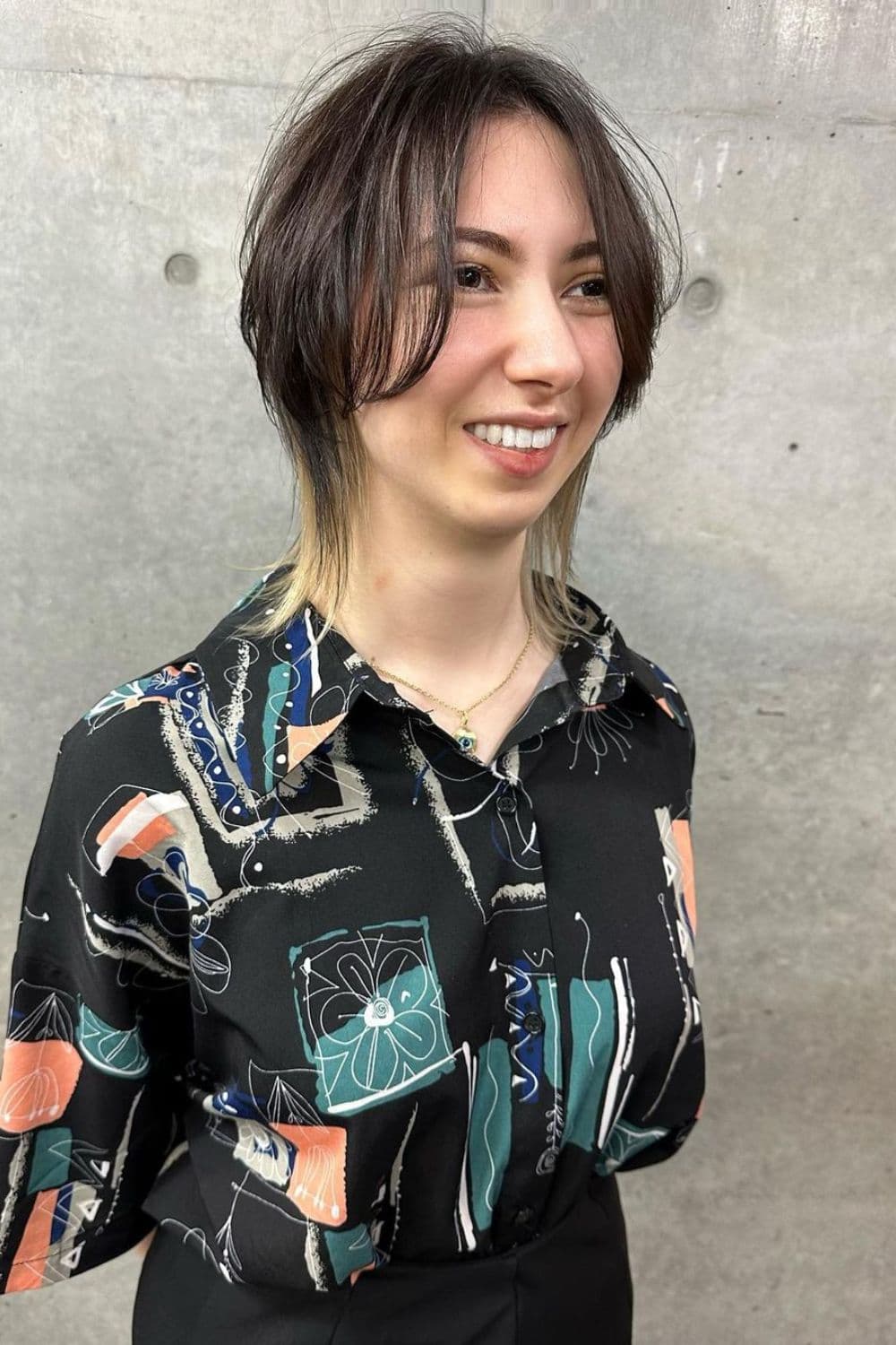 A woman wearing a black buttoned blouse with print and a short black and blonde peekaboo wolf cut.