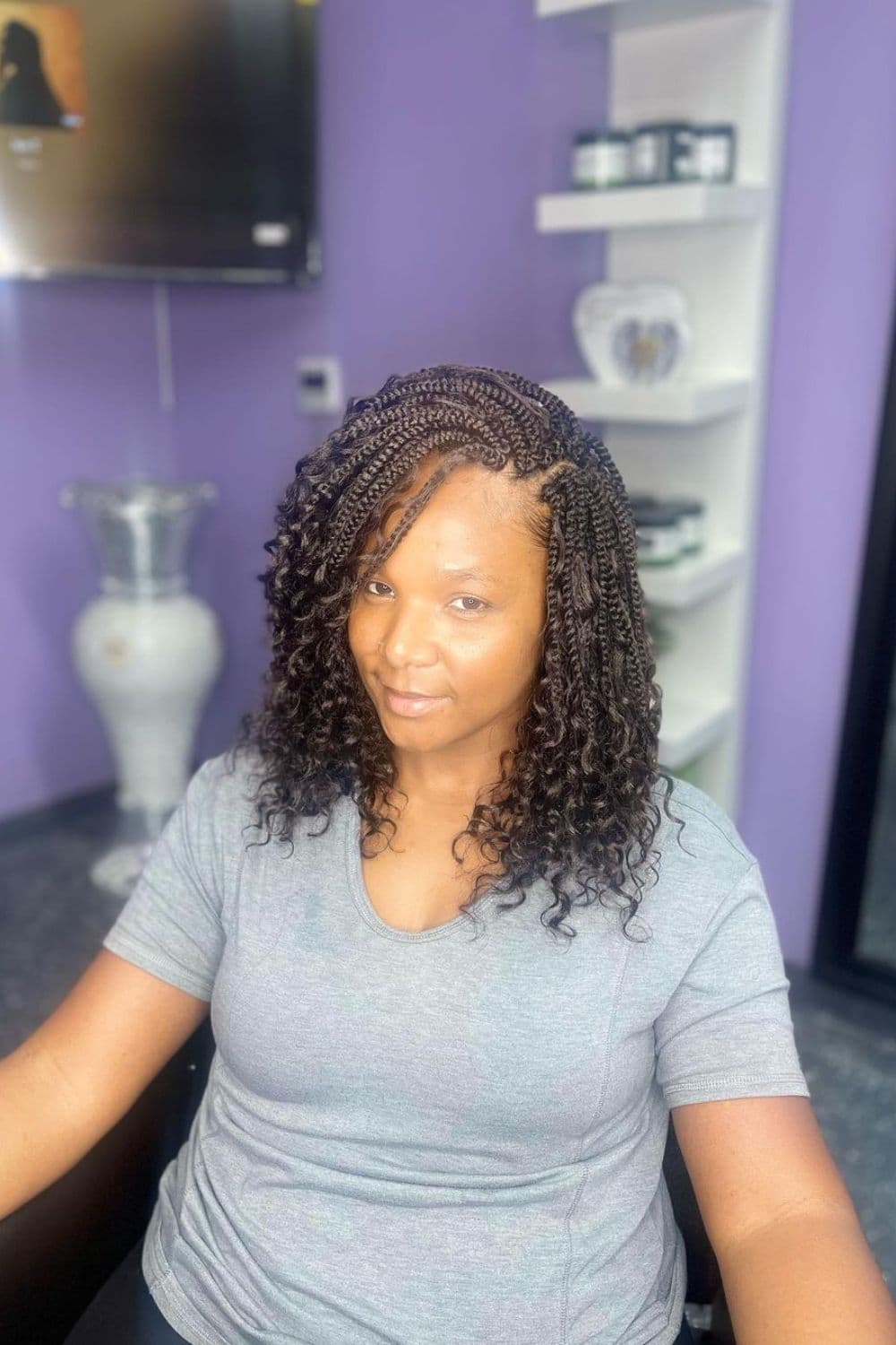 A woman sitting on a chair in the salon with black shoulder-length box braids with curly ends.