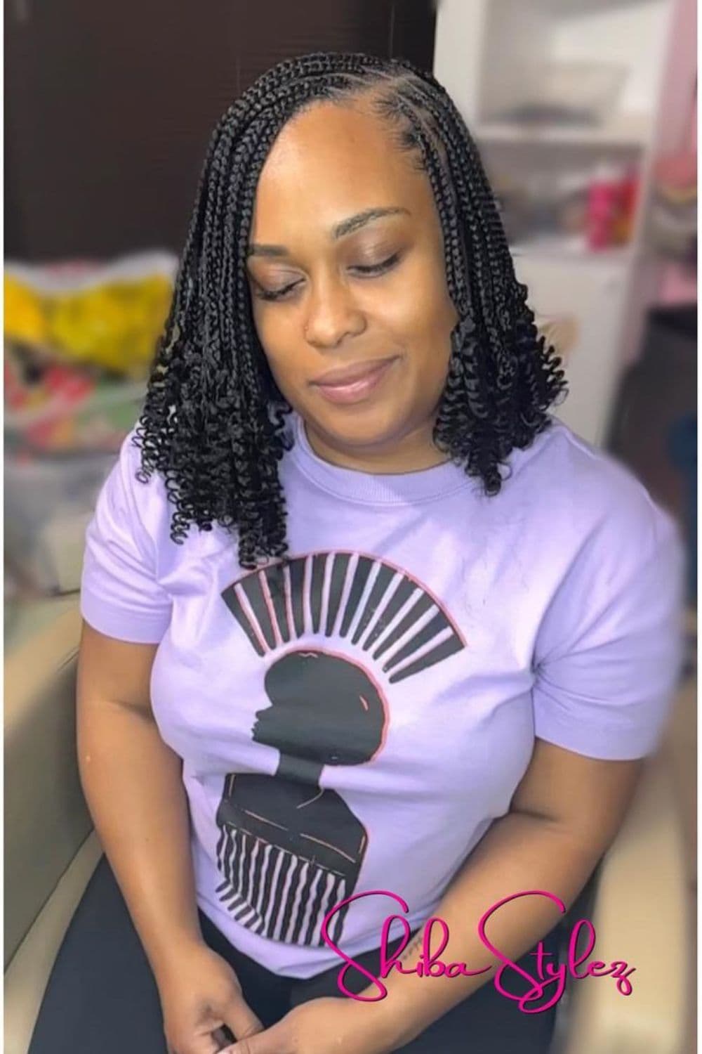 A woman sitting wearing a lavender t-shirt with black shoulder-length medium knotless braids with curly ends.