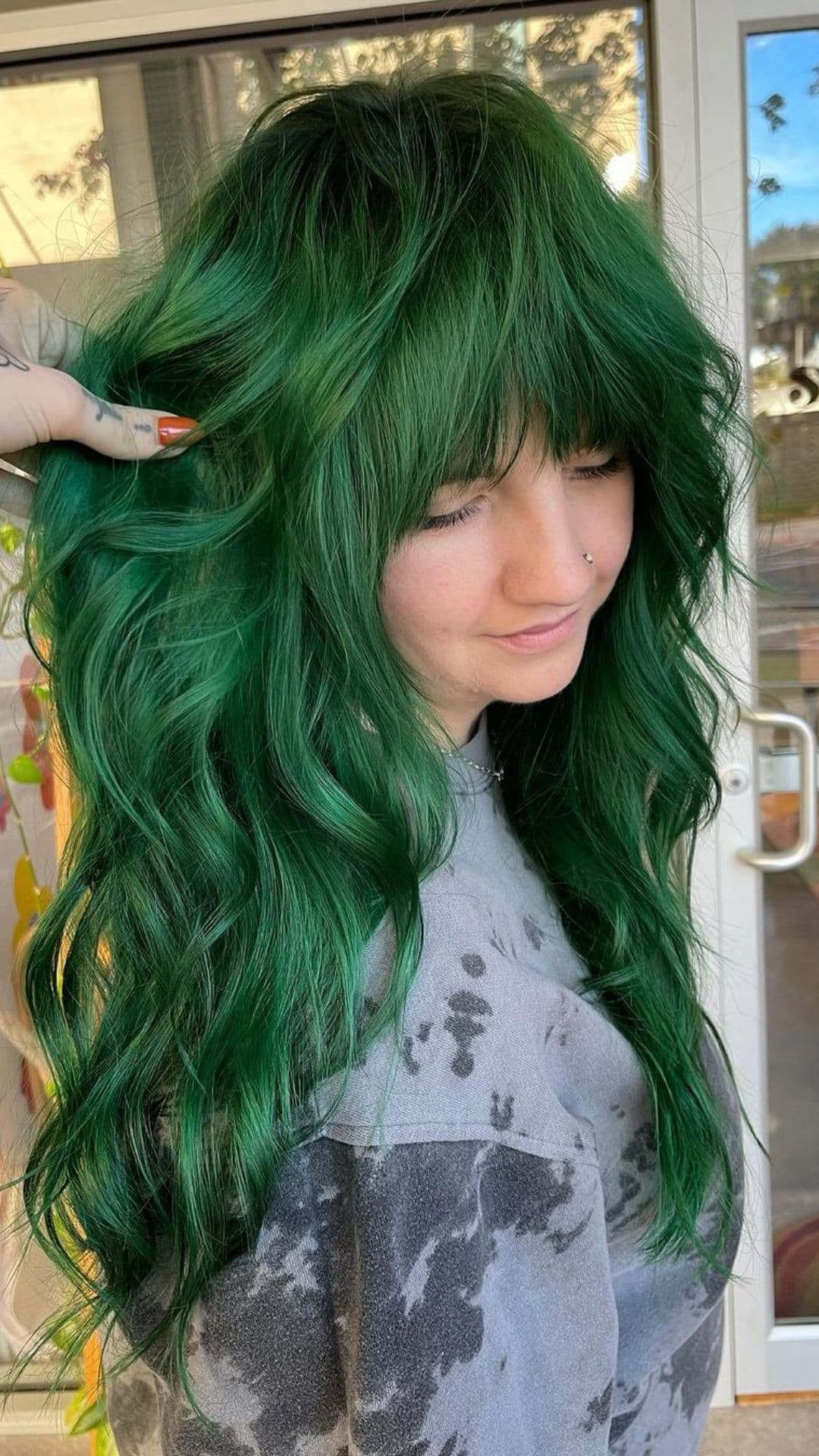 Person with long wavy hair dyed in Christmas Tree Green, wearing a tie-dye sweater, standing indoors.