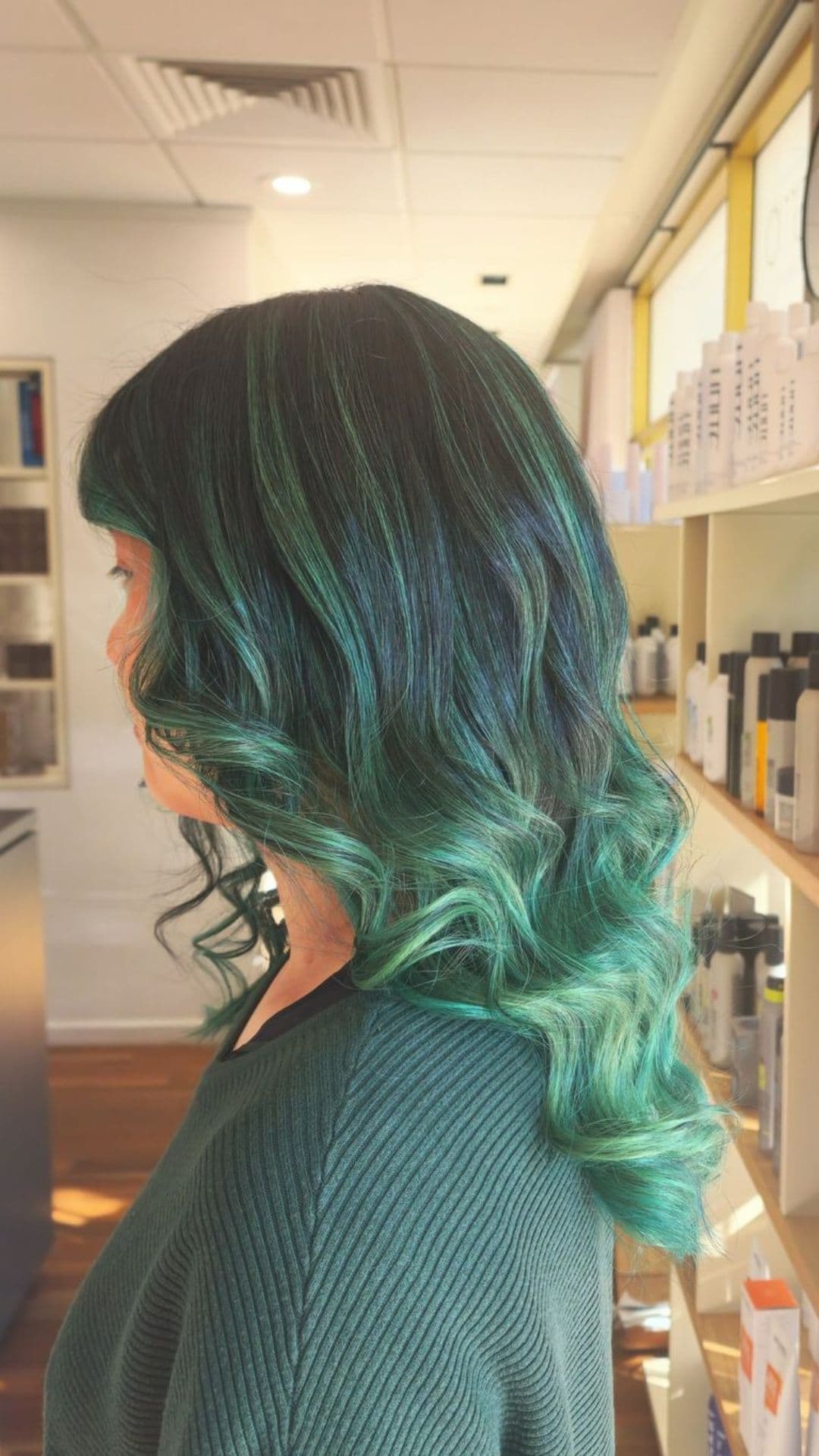 Side view of a person with curled Mistletoe Green Ombre hair, standing in a salon.