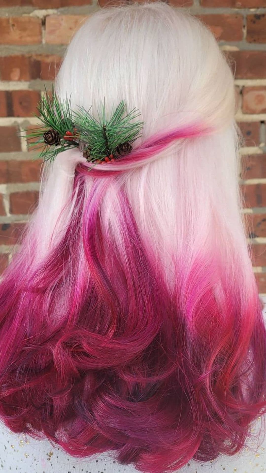 Woman with snow white hair and cranberry tips, styled with a pine and berry hair accessory, in front of a brick wall.