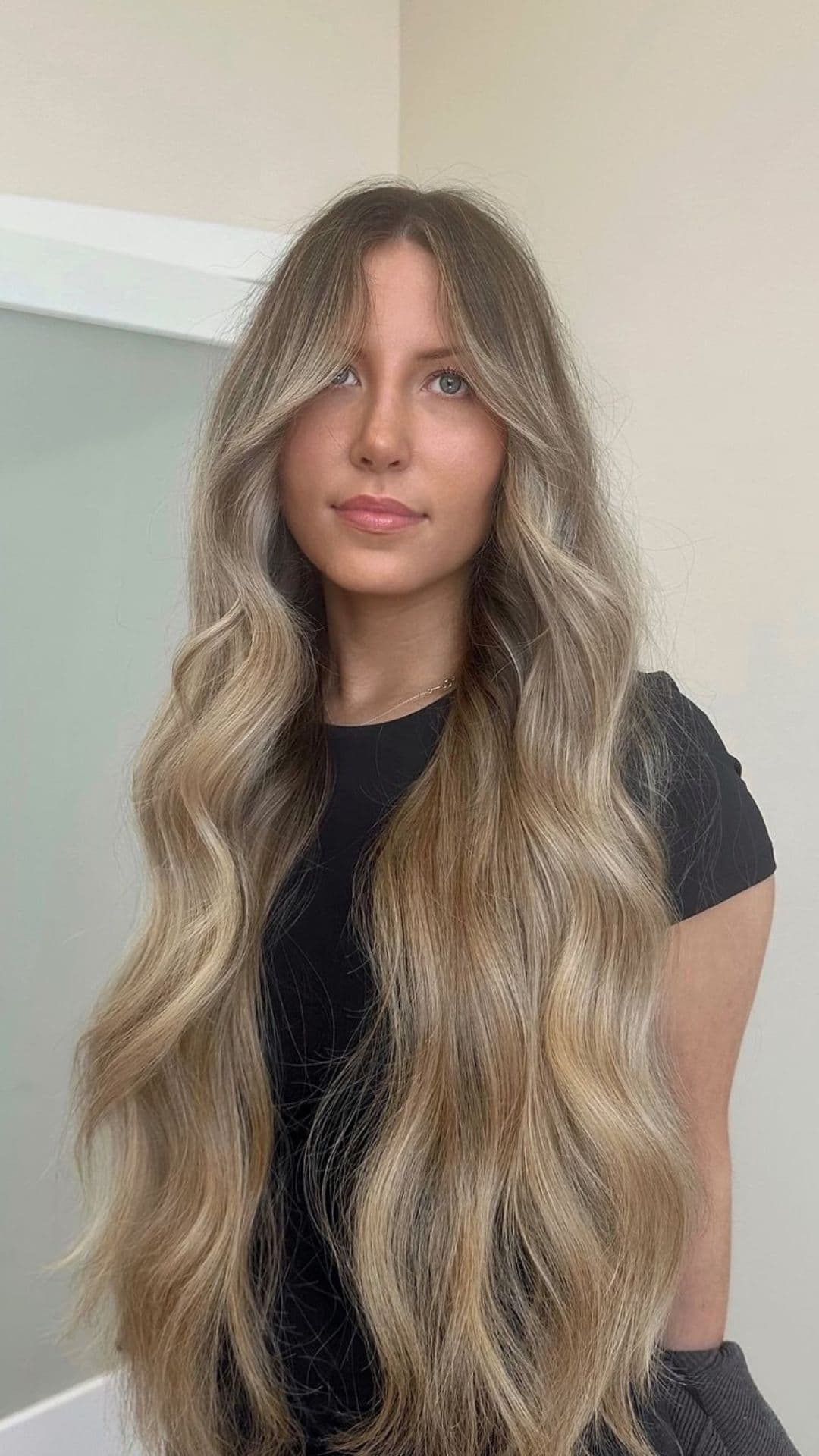 Woman with long wavy Blended Bronde hairstyle, wearing a black top, standing against a light background.