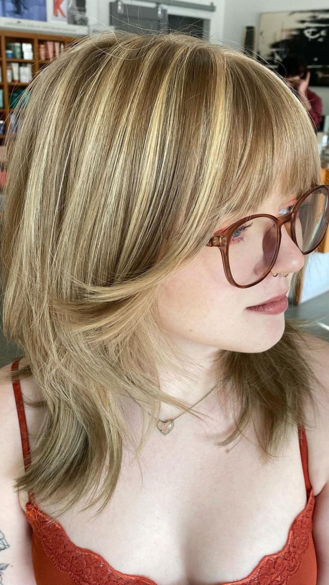 Woman with blonde highlights hairstyle, wearing glasses, orange top, and heart necklace.