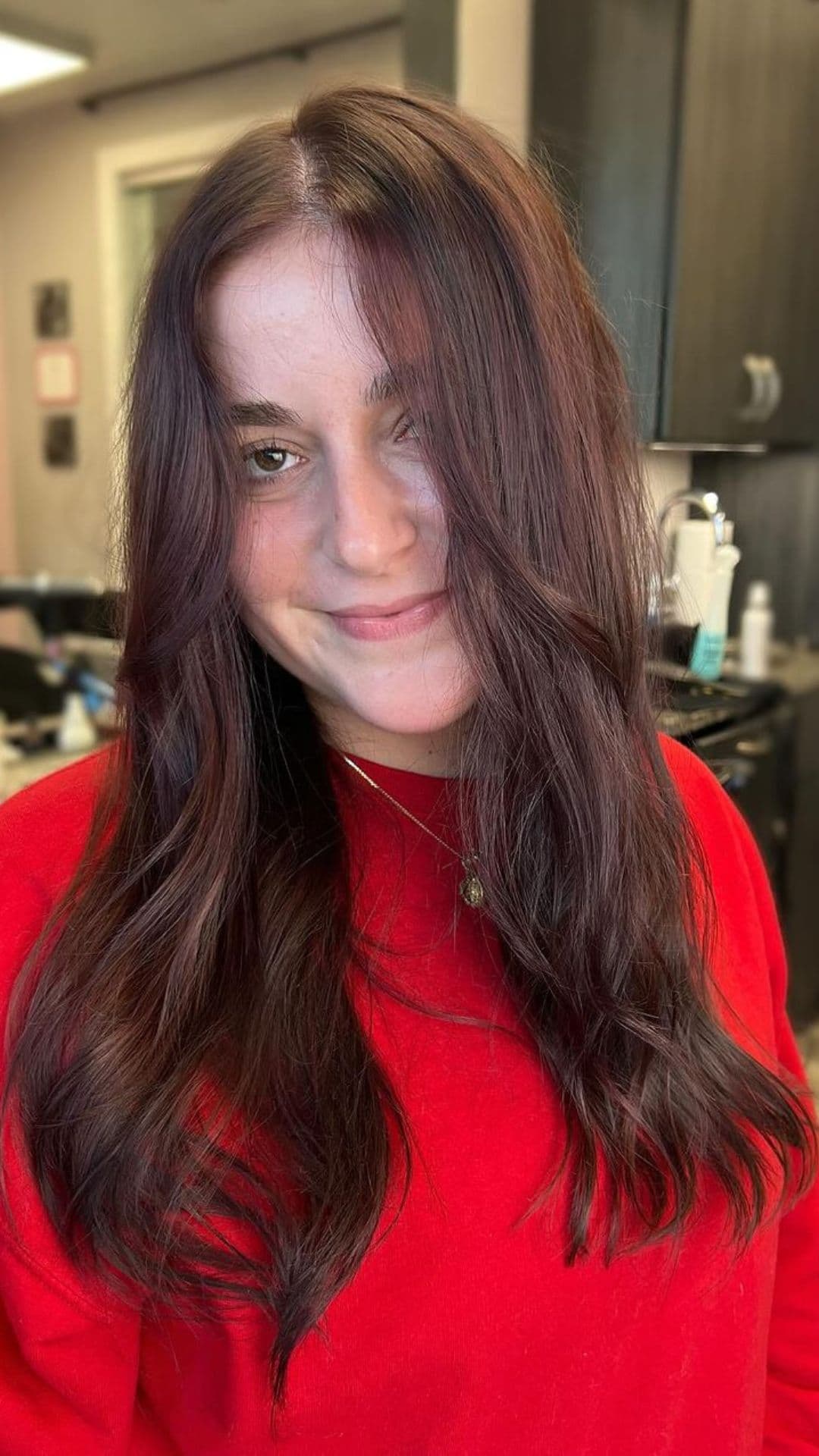 Woman with long, wavy Chocolate Cherry hairstyle, wearing a red sweater, smiling softly at the camera.