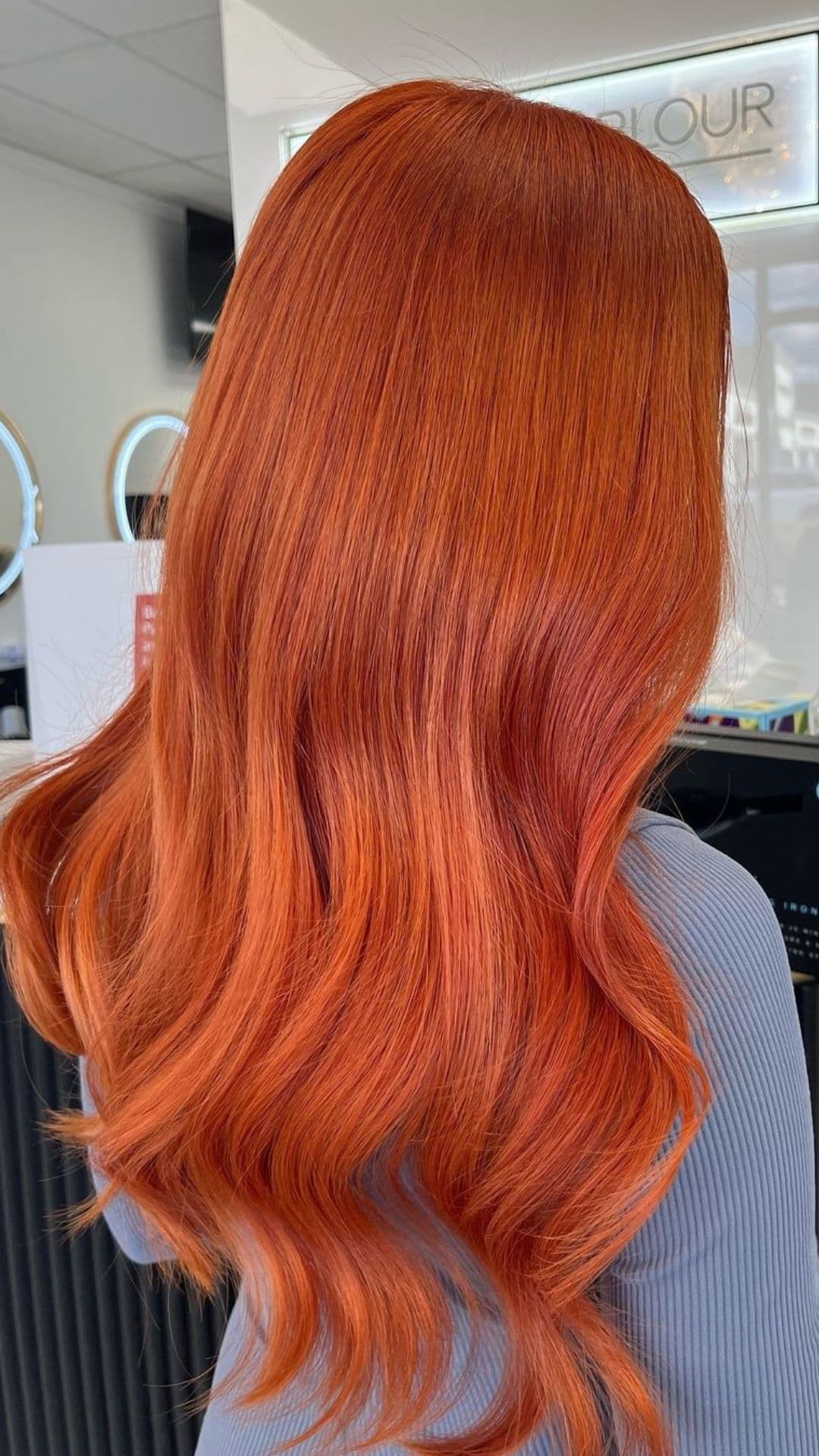 A person with long, wavy copper hair shown from the back in a salon setting.