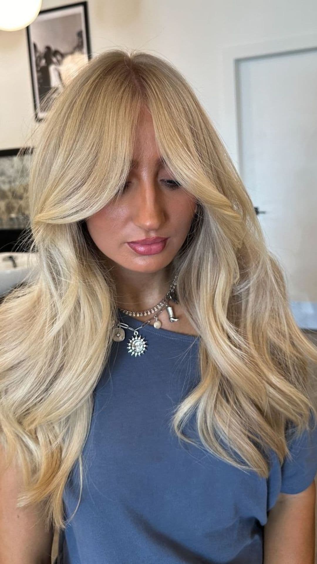 A person models a golden blonde hairstyle with long layered waves. They wear a blue top and a silver necklace.