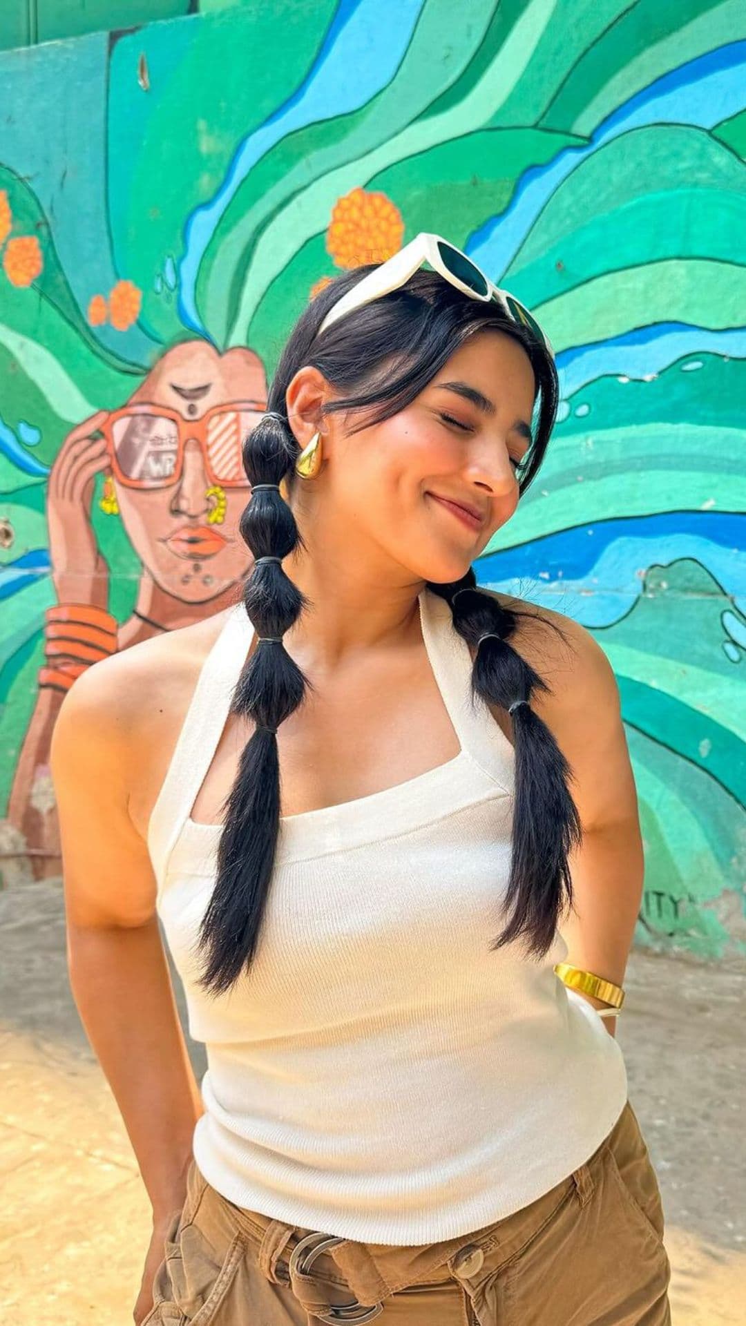 Woman with Bubble Braids hairstyle, wearing a white top and accessorizing with sunglasses and earrings, stands against a colorful mural background.