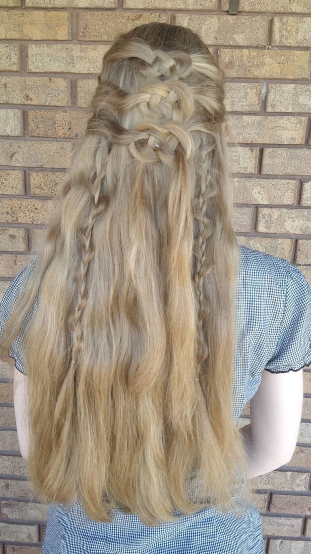 Back view of a person with long blonde hair styled in a Celtic Knot. Brick wall in the background.