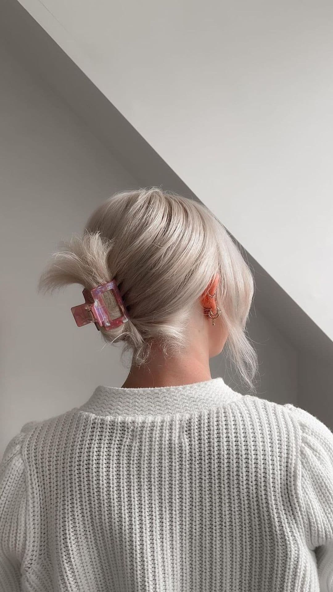 Woman with ash blonde hair shows off a Claw Clips Up-Do hairstyle. She uses a pink claw clip to secure her hair in a neat up-do. She wears a white knit sweater. The setting appears minimal and well-lit.