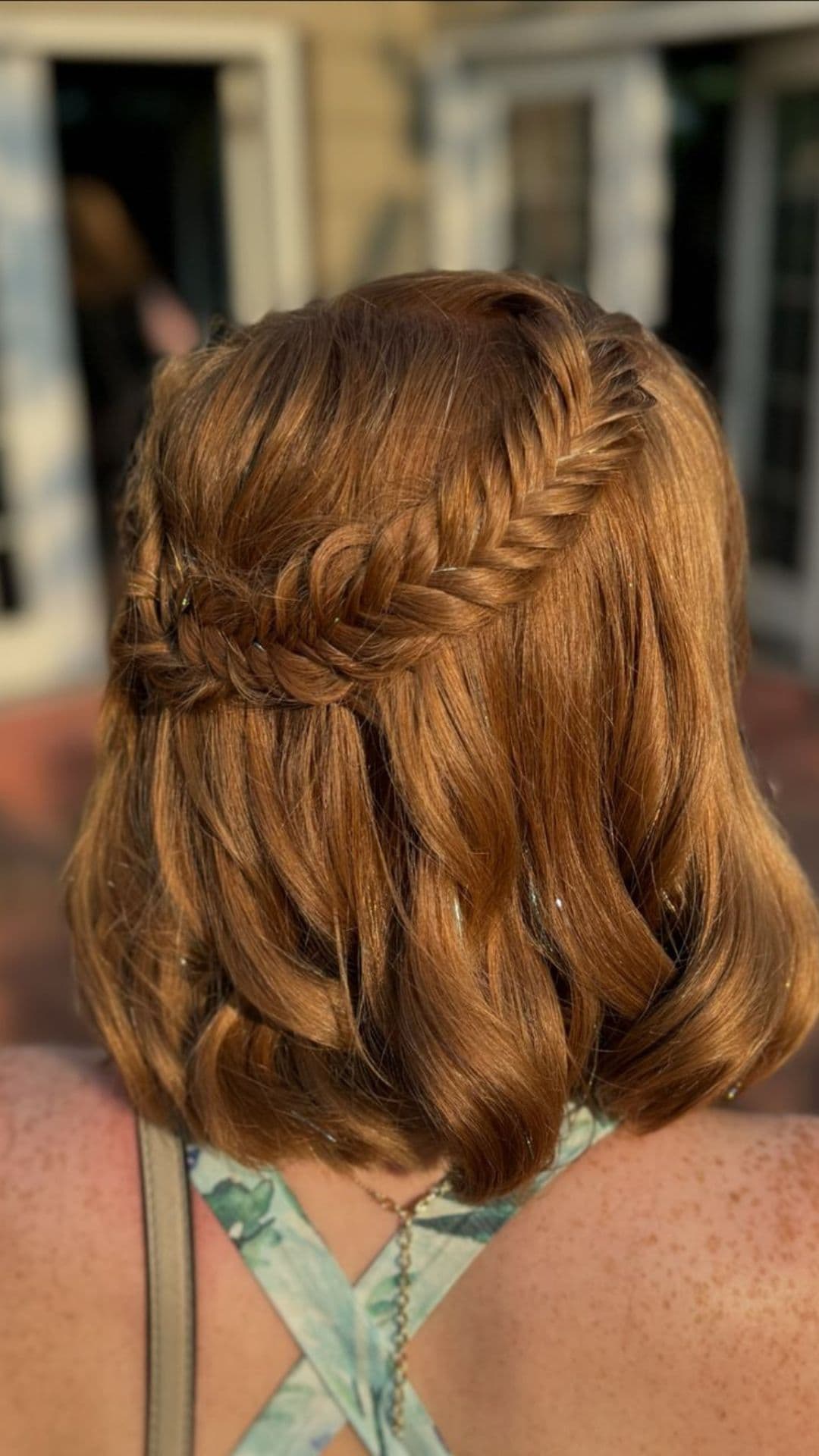 Back view of a person with shoulder-length red hair styled in a Fishtails Half Updo. They're wearing a strappy top in a sunlit outdoor setting.