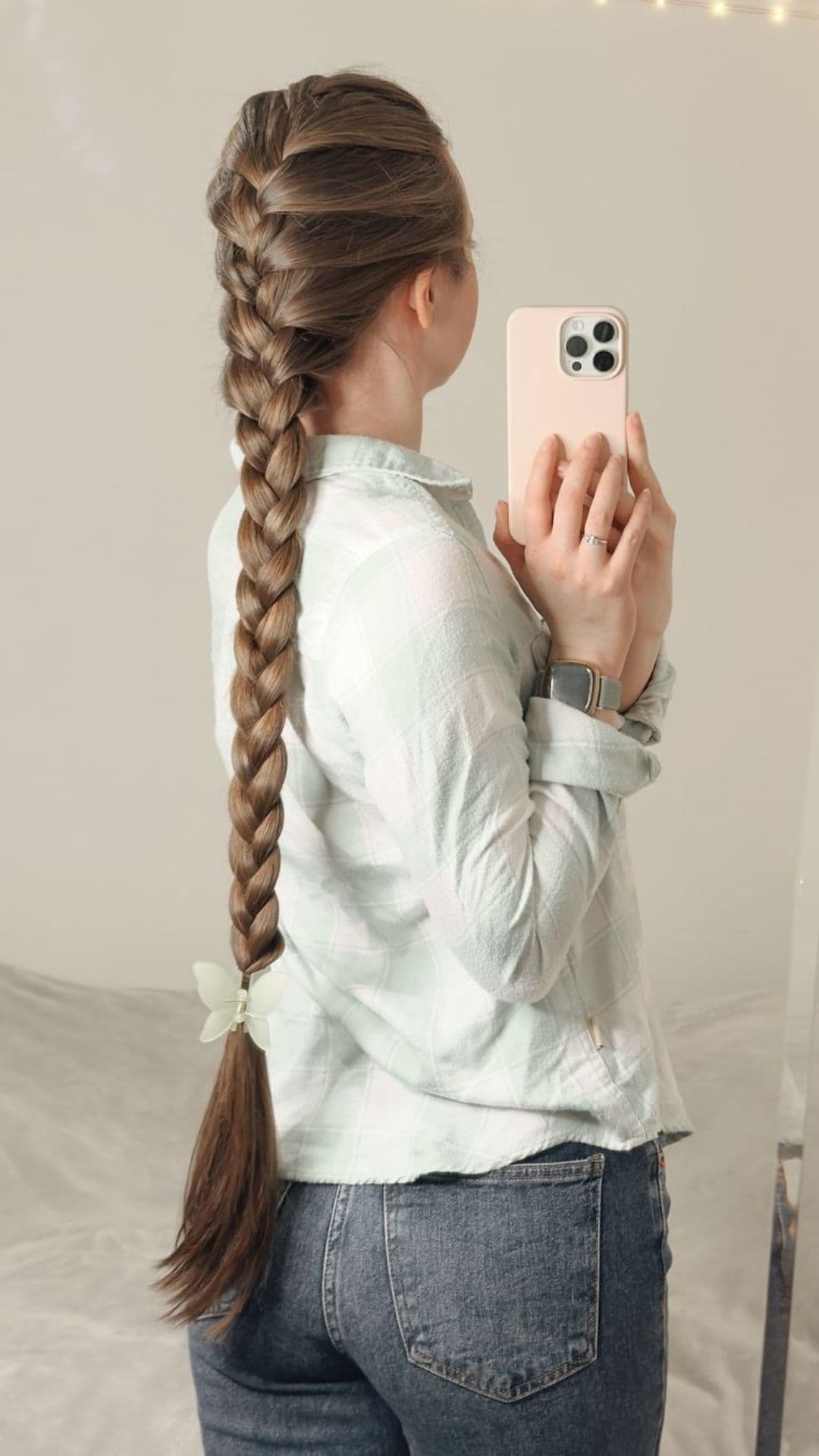 A person modeling a long French braid ponytail from the back, holding a smartphone, wearing a light plaid shirt and jeans.