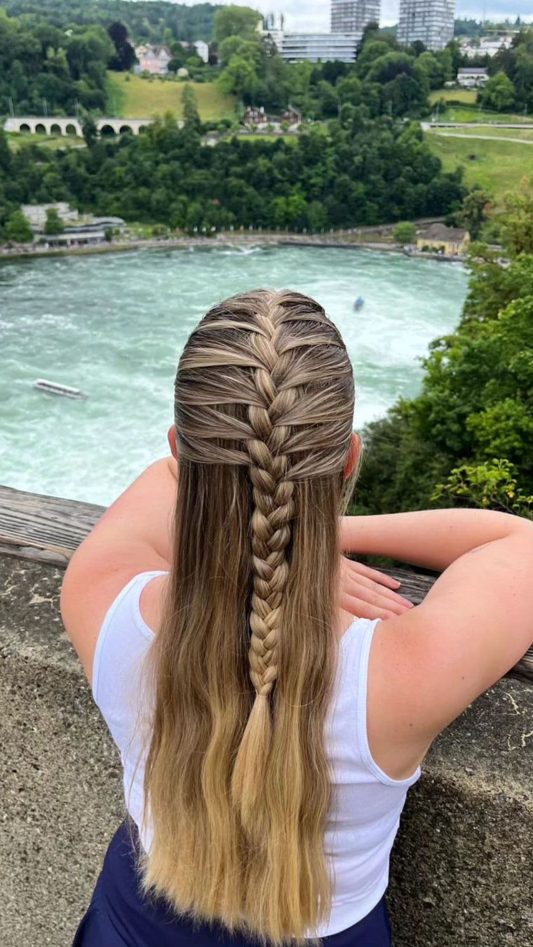 You see the back of a person with long, blonde hair styled in a Half-Up Braid. They're wearing a white tank top, facing a scenic view of a river and greenery.