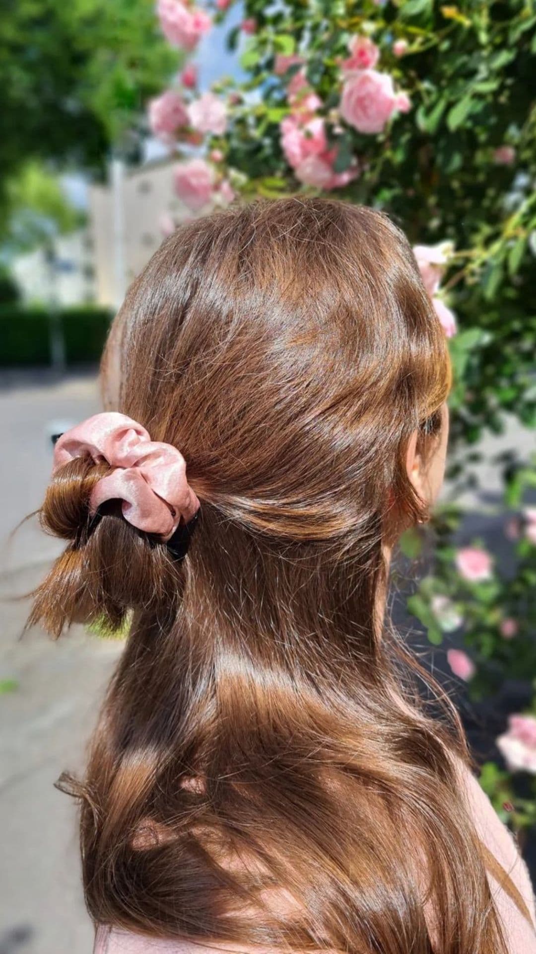 Woman modeling a Half Up Half Down Messy Bun hairstyle with a pink scrunchie, standing near blooming roses.