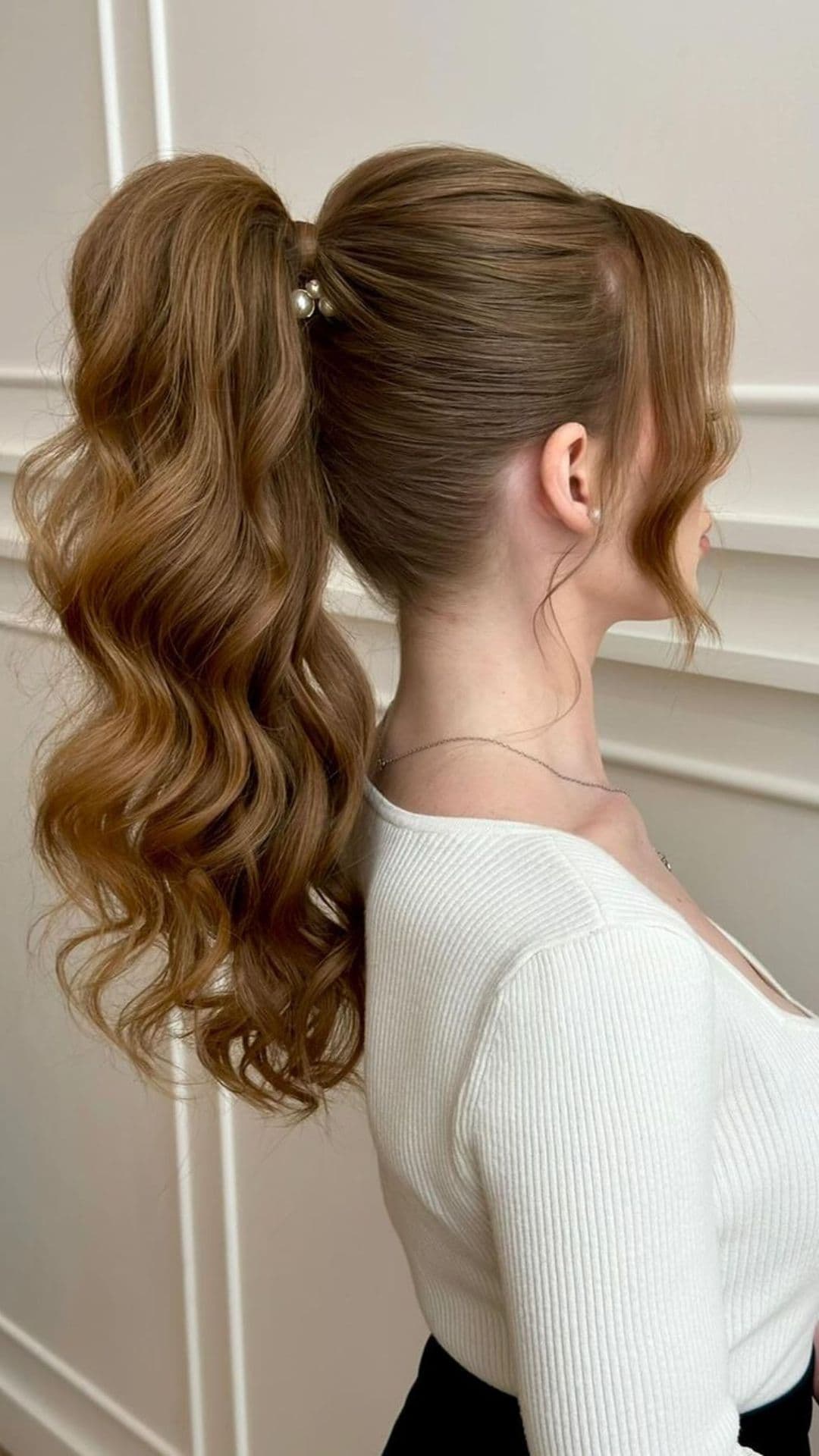 Person modeling a messy high ponytail hairstyle. Hair is long, wavy, and tied with pearl accessories. Wearing a white ribbed top.