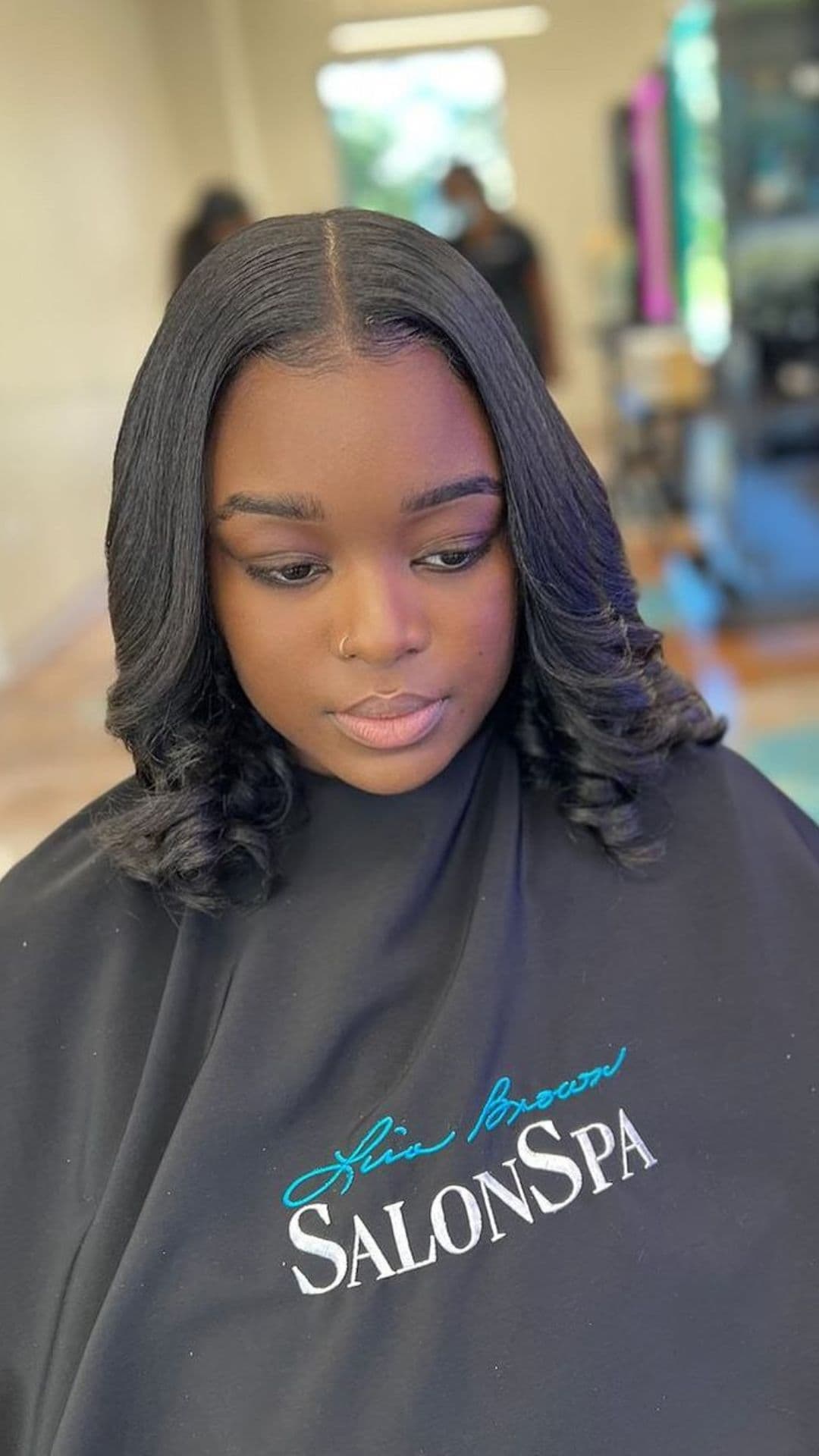 A person with a Silk Press hairstyle and loose curls sits in a salon chair, covered by a black salon cape with 