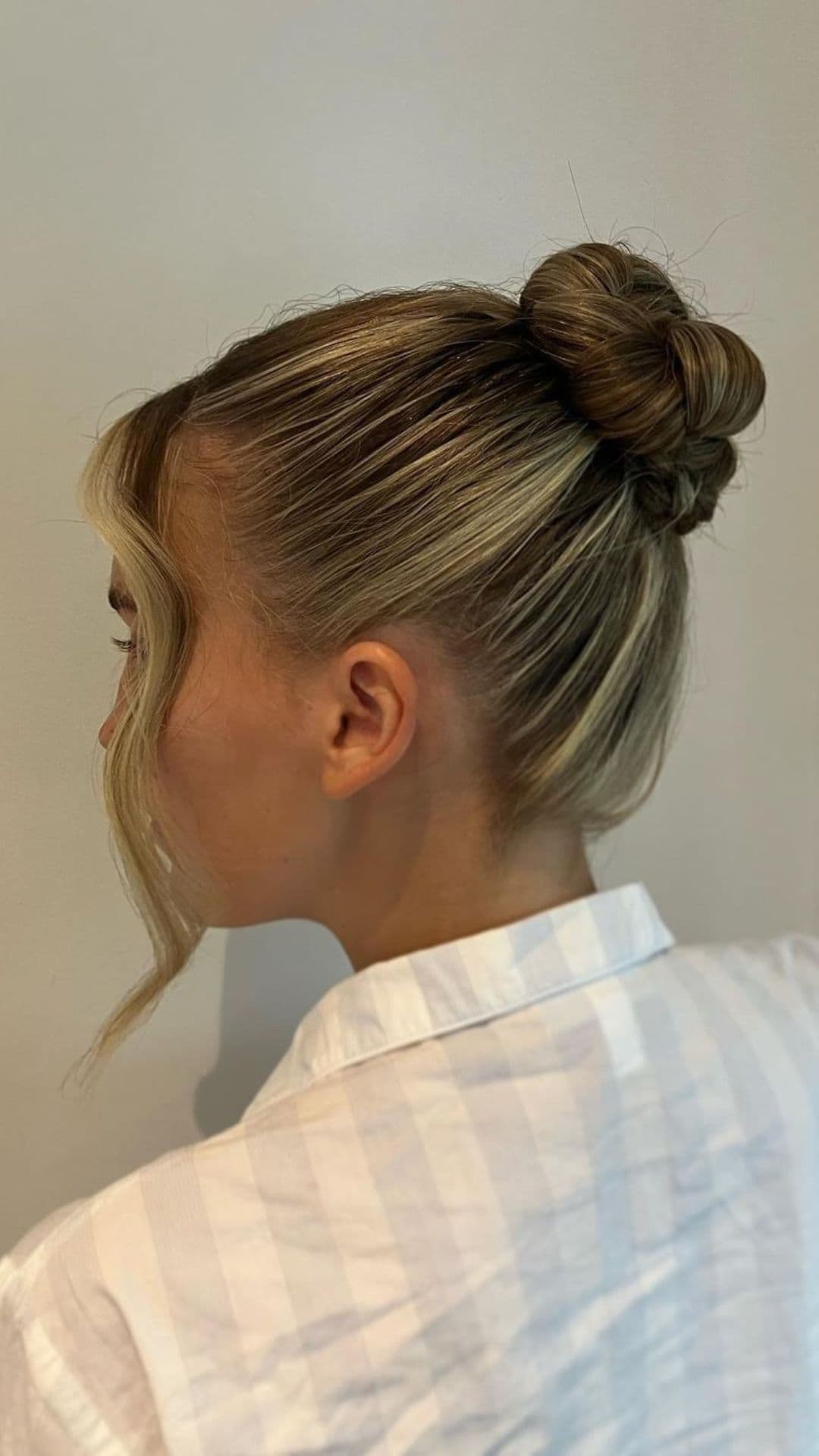 Woman with blonde hair modeling a Twisted Ballerina Bun hairstyle, featuring a smooth updo with a twisted bun at the crown and a loose, curled strand framing the face.