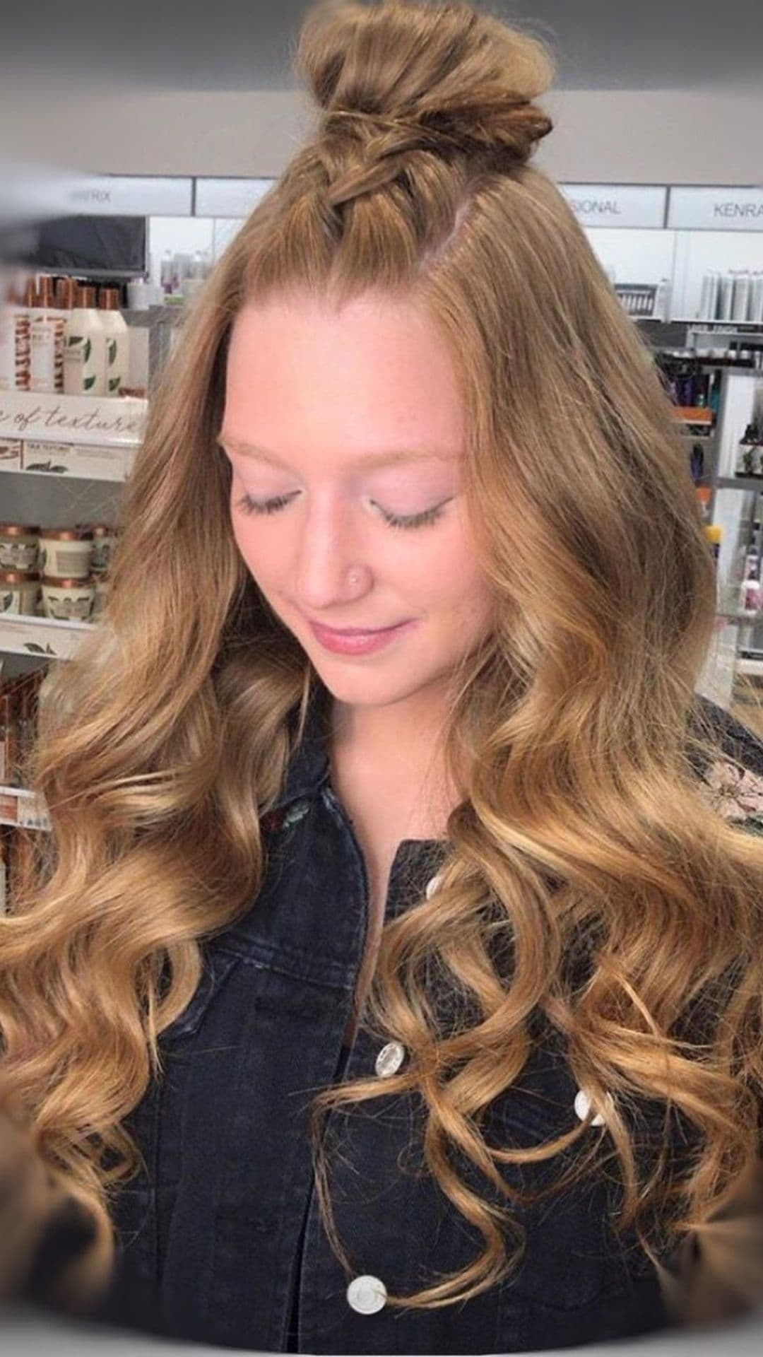 Person with long, wavy braided topknot hairstyle. They're wearing a dark denim jacket. They're smiling gently with closed eyes. The background features shelves with beauty products.