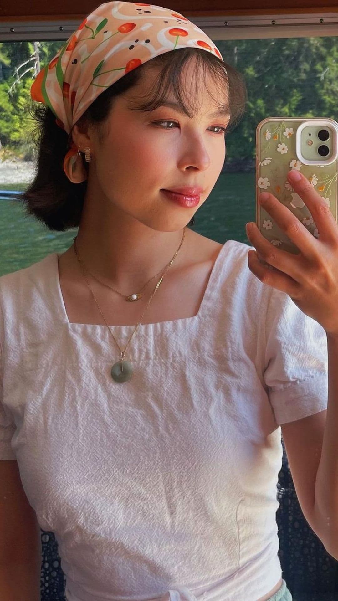 A person taking a selfie in front of a window. They wear a vibrant bandana and a simple white top. Their hair is styled in a Bandana Hairstyle.