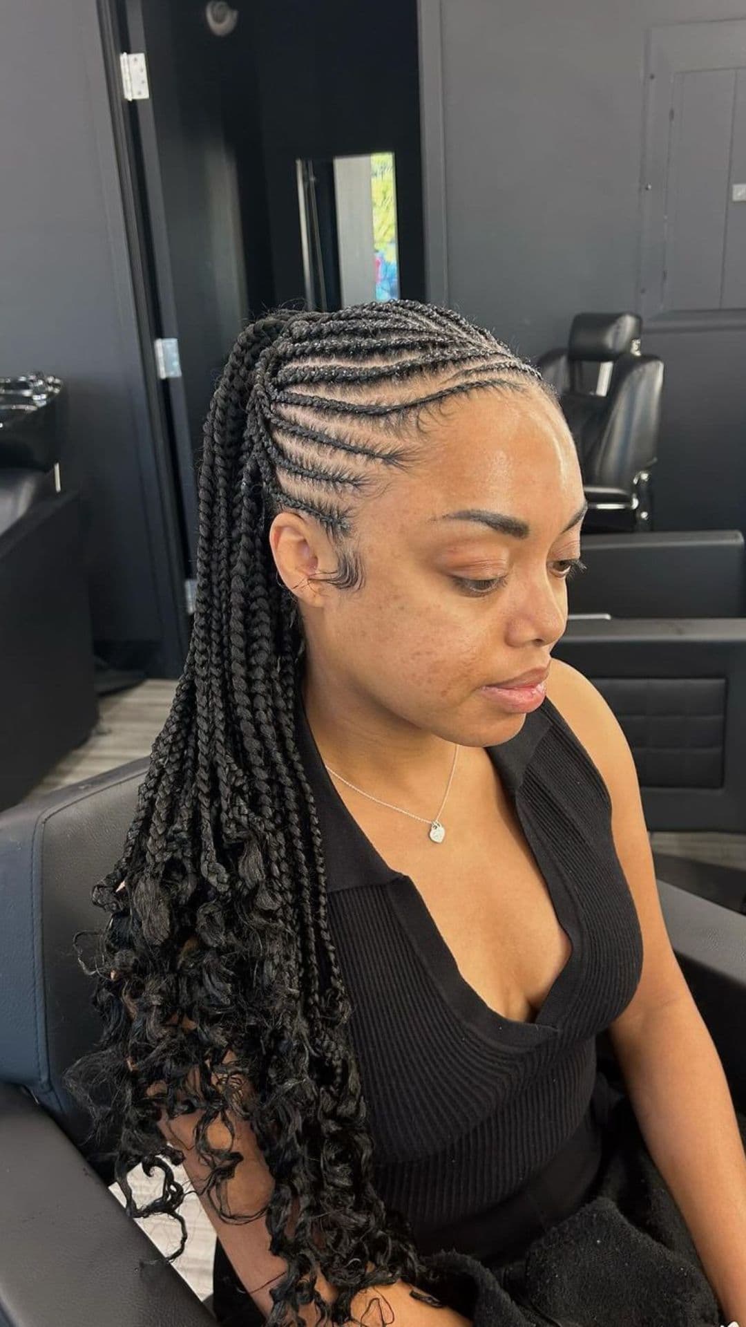 A woman sitting in a salon chair, modeling cornrows with long, curly braids.