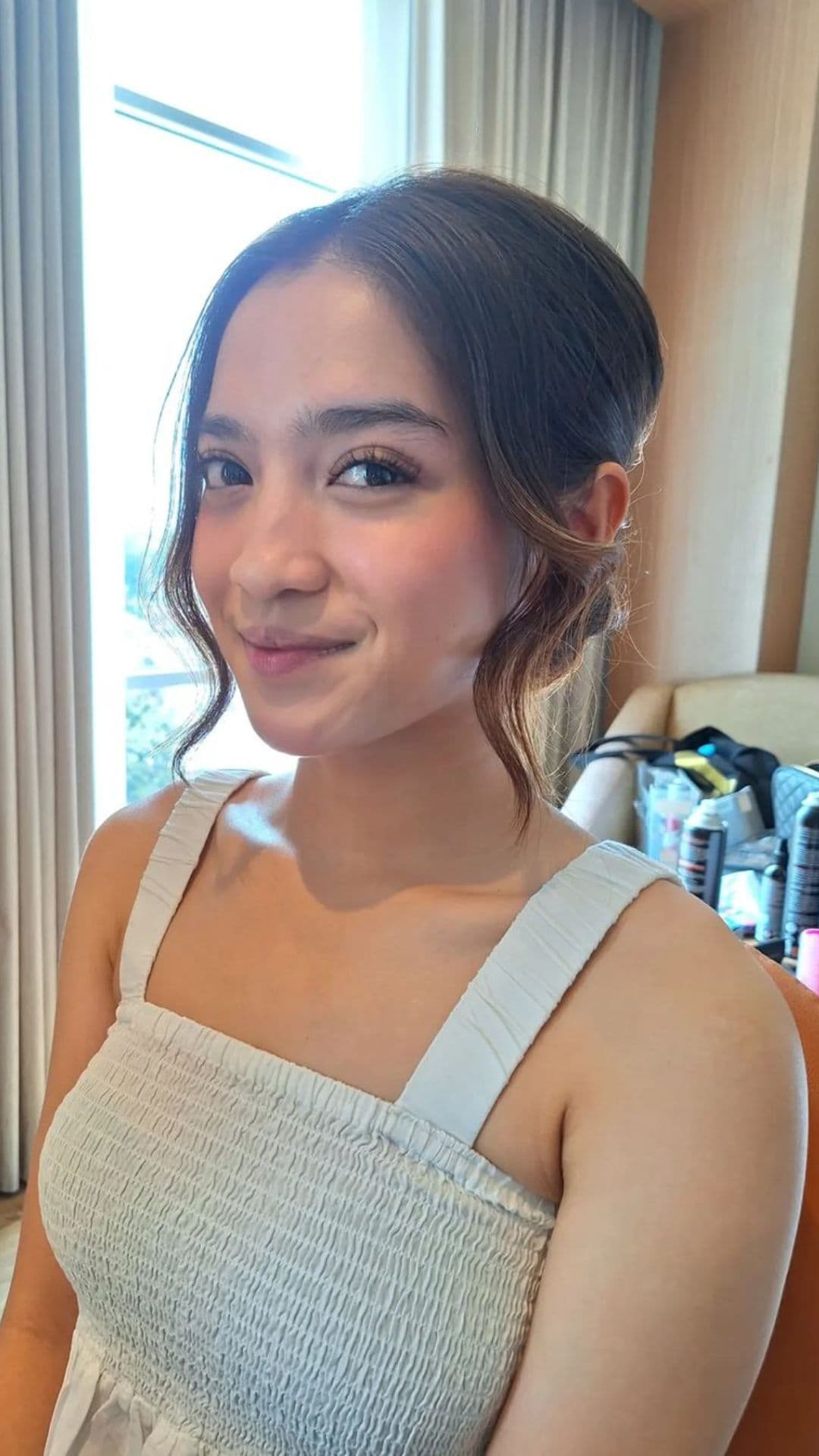 A person models a low bun with a middle part hairstyle, wearing a sleeveless white dress and smiling softly. The background shows a window and a dressing table with hair products.