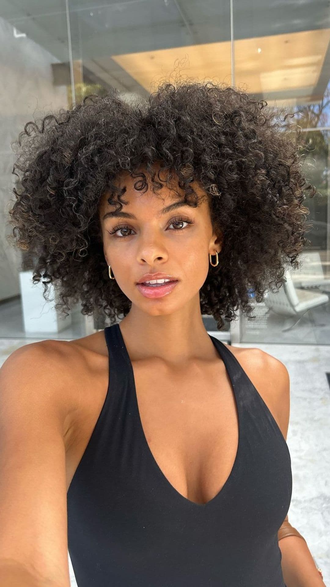 A person with natural curly hair takes a selfie outside. They wear a black top and gold hoop earrings.