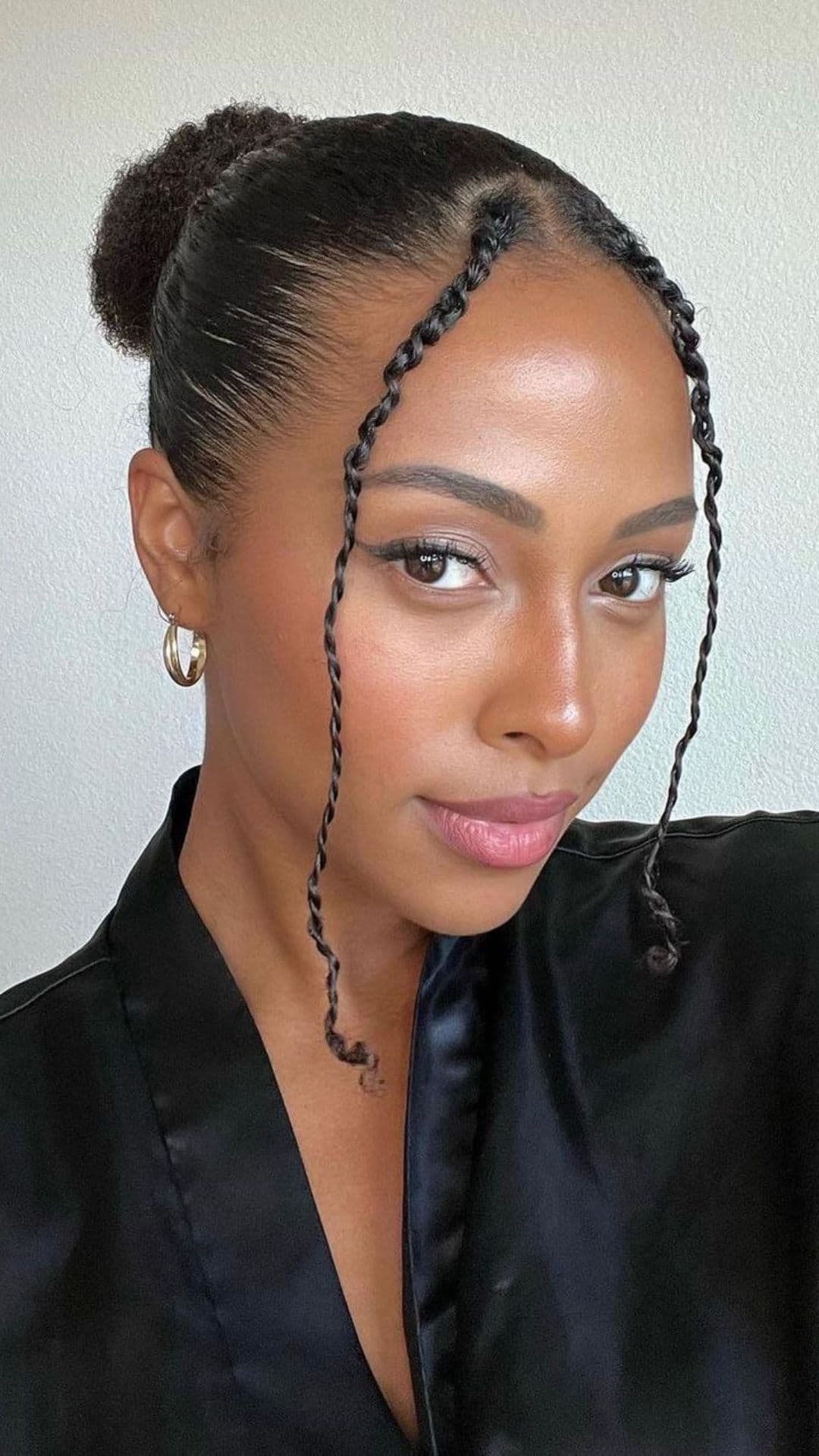 A person modeling a slicked bun hairstyle with two twisted tendrils framing the face, wearing gold hoop earrings and a black satin top.