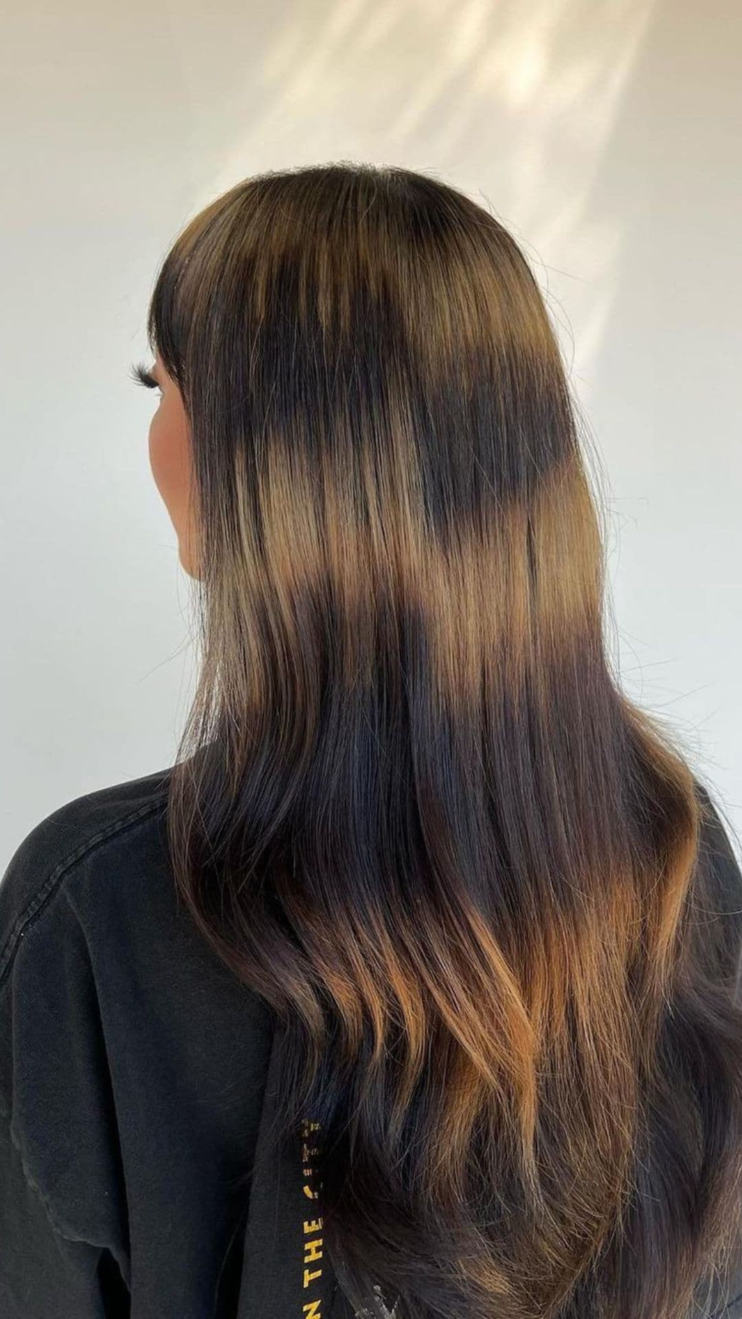 Woman modeling Tortoiseshell Brunette hairstyle with long, flowing hair featuring dark brown and caramel tones. She's wearing a black shirt, looking away from the camera.